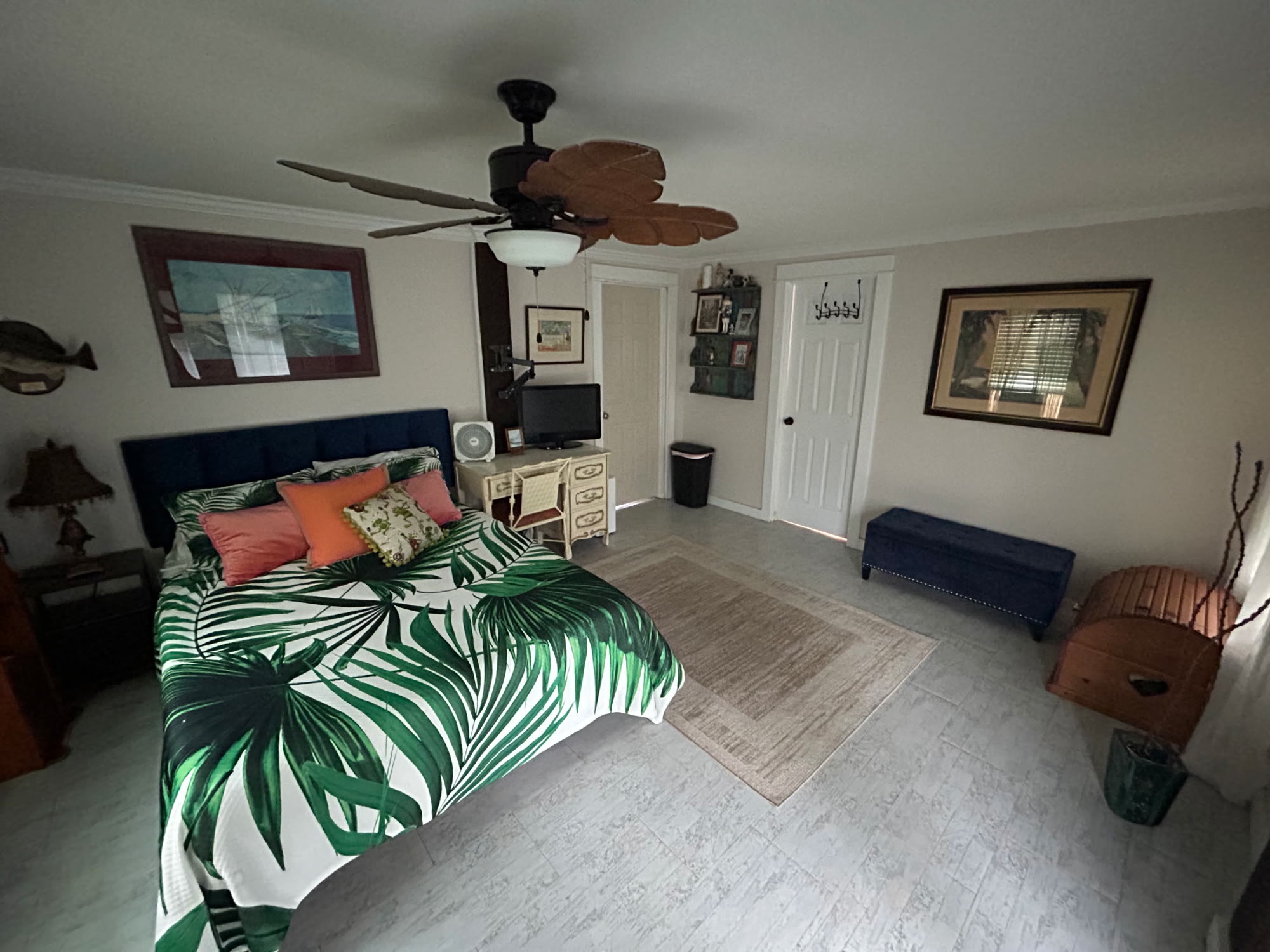 Guest bedroom with a navy upholstered headboard, palm-print bedding, coastal artwork, and a ceiling fan