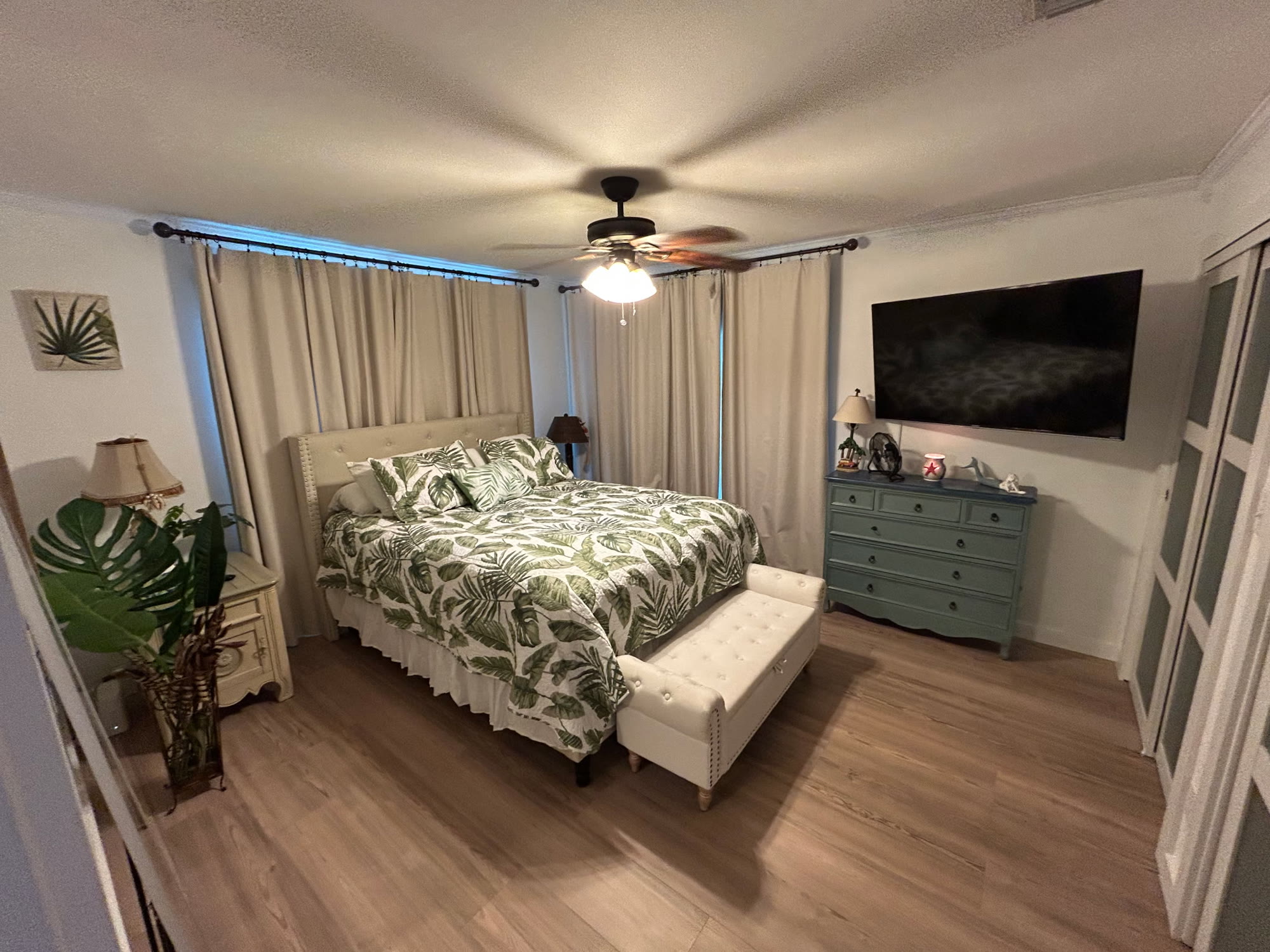 Master bedroom with a king bed in palm-print bedding, upholstered headboard, teal dresser, and wall-mounted TV